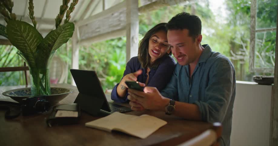 Young couple talk while browsing on a smartphone sitting at a table at home