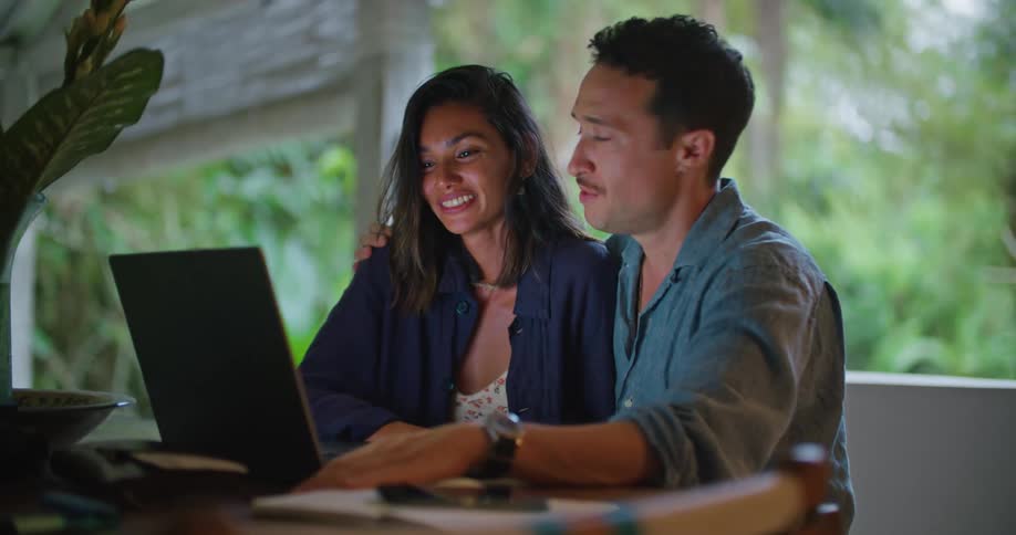 A man and woman sit at the kitchen table looking at something on a laptop while laughing and being flirty