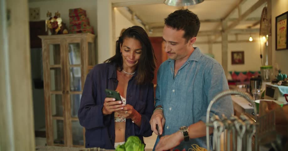 Husband cuts vegetables at the kitchen counter while wife shows him something on her phone and they both laugh