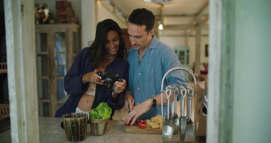 A woman share photos on her camera with her date who is cutting and slicing on a cutting board
