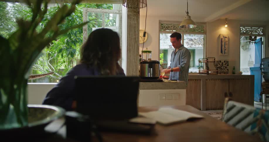 Husband cooks dinner in a kitchen while wife works on a laptop at the kitchen table