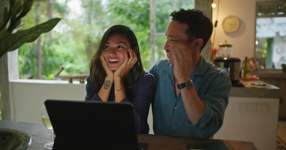Young couple sitting at the dinner table hug and laugh while looking at a laptop