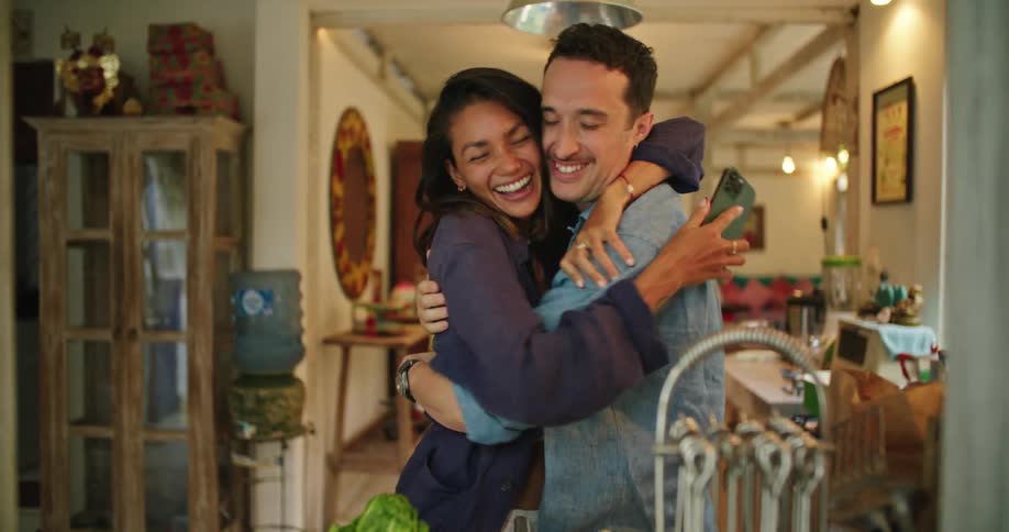 Husband and wife are in their kitchen hugging and laughing after looking at something on a smartphone