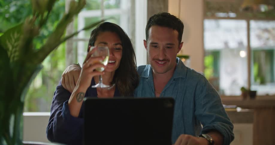 A husband pours wine for his wife and walks over to her where she is looking at something on a laptop