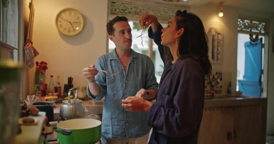 A man and a woman are standing in a kitchen as he is cooking on the stove he takes a noodle out of the pot for her to taste