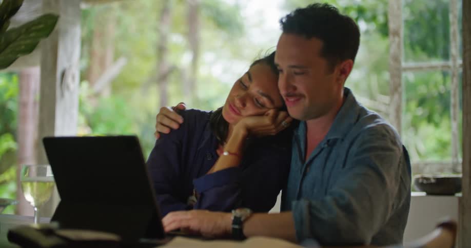 A man and a woman are sitting together at a table with a laptop computer talking and he hugs and kisses her