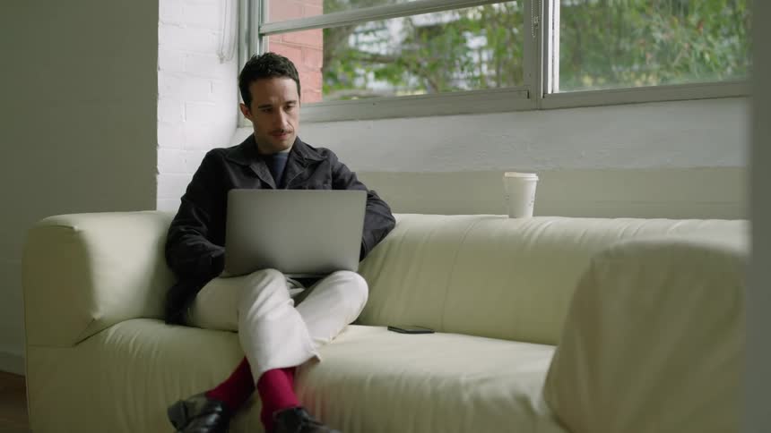 A man sits on his couch with a laptop on his lap sipping coffee and working on something