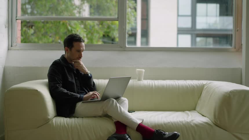 Man typing while sitting on couch takes break to drink from portable cup