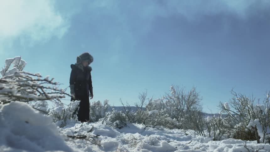 A man in heavy clothing stands in snow and looks downward
