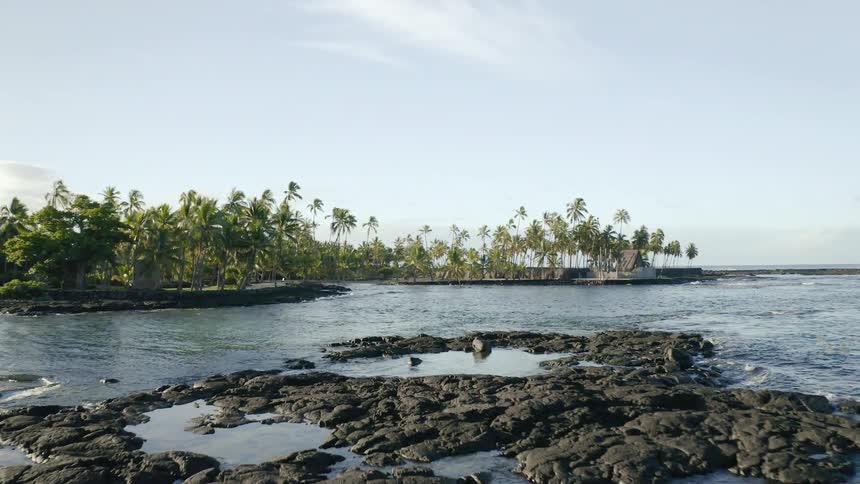Ocean waves lap against the palm tree covered shore and over natural rock pools