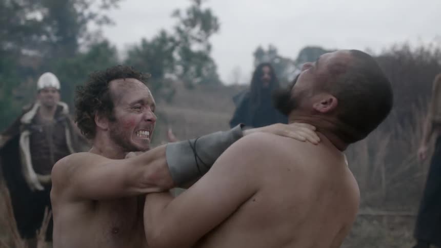 Topless men wrestling in the woods with an audience
