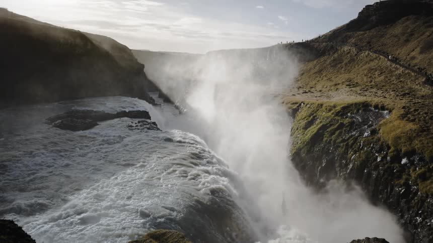 People stand along a trail and watch as waterfall fills river valley with mist