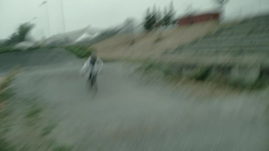 Cyclist rides bike over hilly path passing audience stand and toward large homes