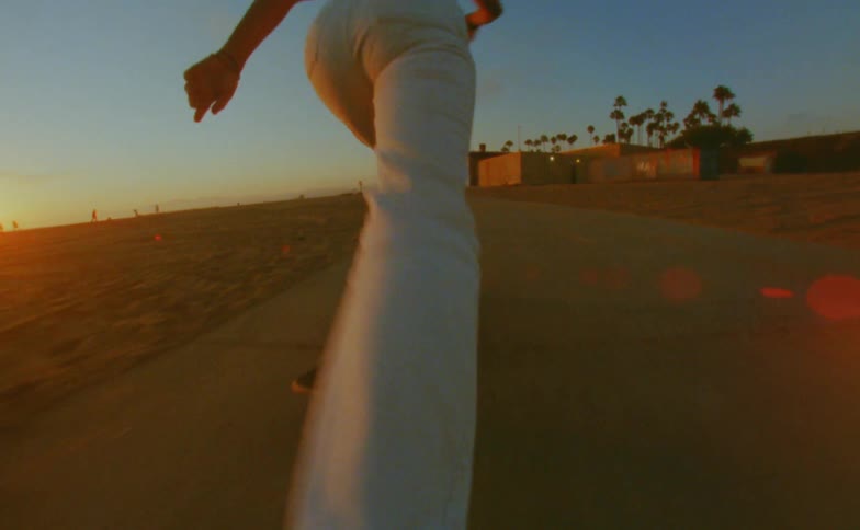 Woman with cap yellow top and white pants skateboards on road on the beach at sunset
