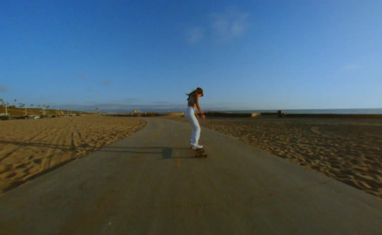 Woman with cap yellow top and white pants skateboards on road on the beach