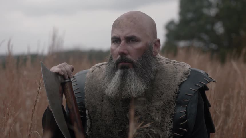 A man dressed as a warrior and holding an axe while standing in some tall grass on a cloudy day
