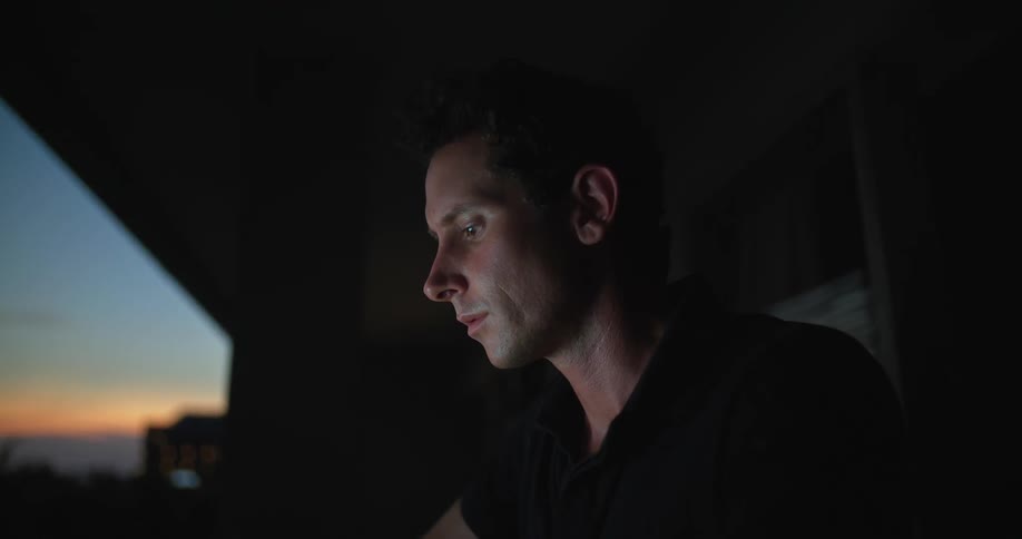 A man is sitting at a table in front of a window typing on a laptop computer