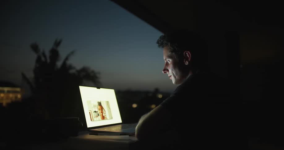 A man is sitting at a desk in front of a window having a video conversation with a woman on a laptop computer