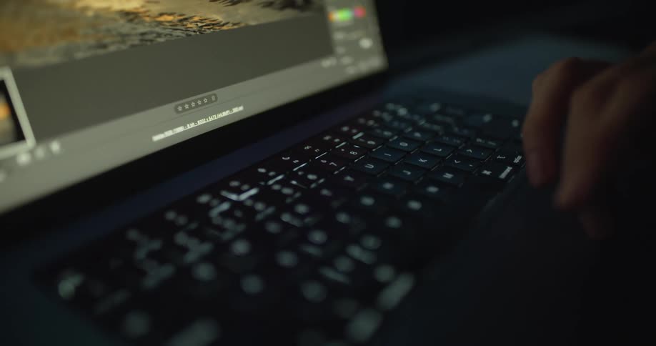 Someone is sitting at a table in a dark room typing on a laptop computer and using the touch pad