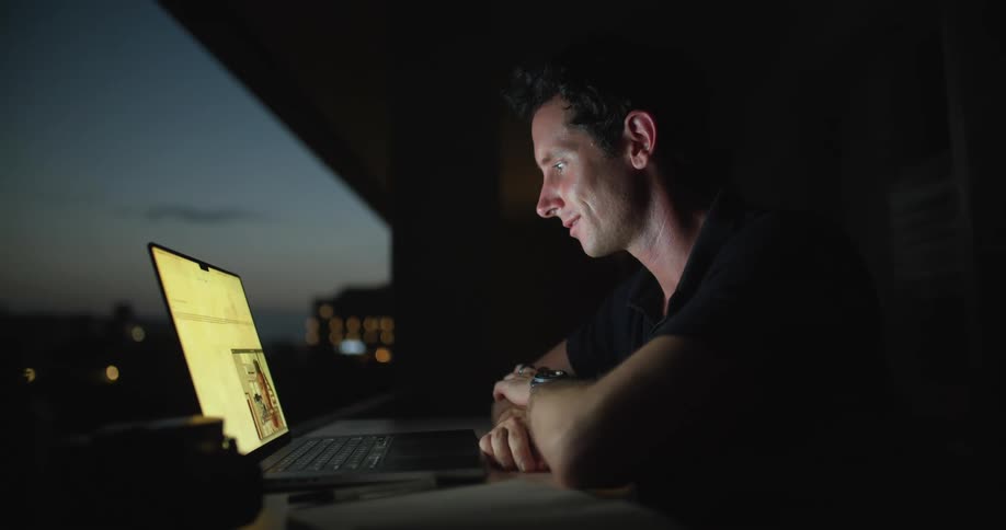 Man sitting at the desk on his balcony having a video call on his laptop at night