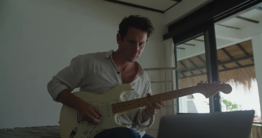 A man is sitting down playing his electric guitar