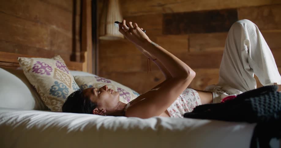Sunlight shines upon a woman lying on a bed and holding her phone in the air as she texts