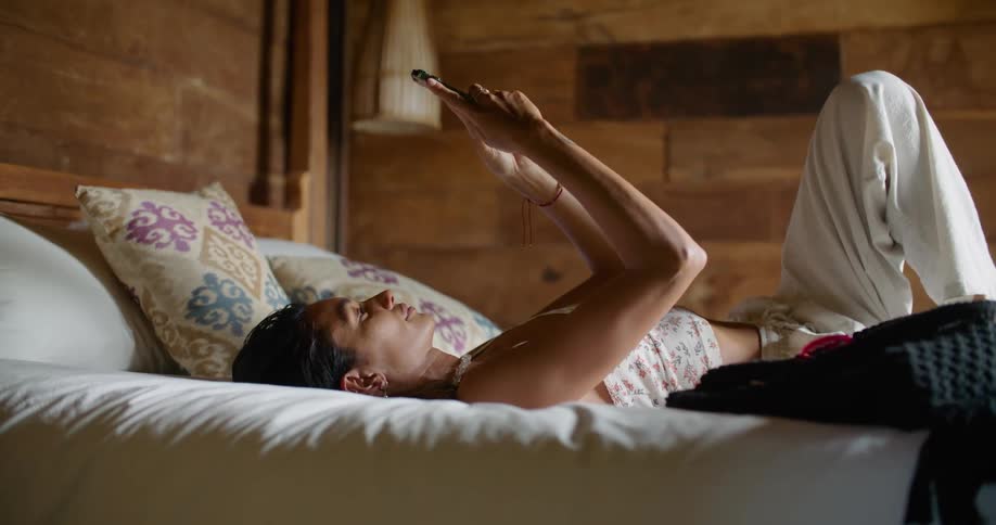 Woman with white top and pants lies on bed using tablet and smiling slightly in bedroom
