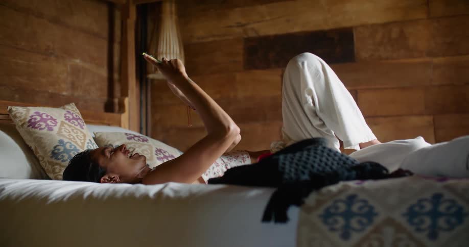 A woman lays on a bed on her back holding up a smartphone while talking and laughing