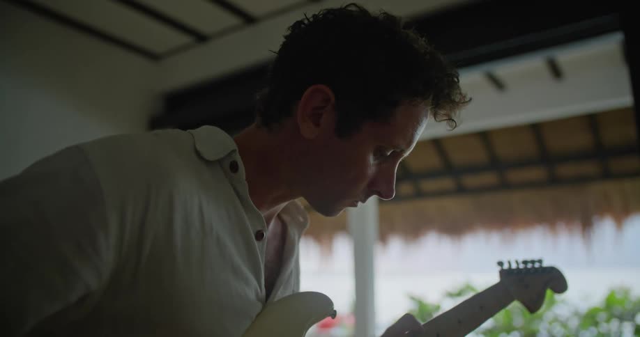 Man with white polo shirt plays white electric guitar sitting on bed