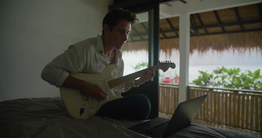Young man sitting in the bed of a hotel room plays electric guitar while looking at a laptop