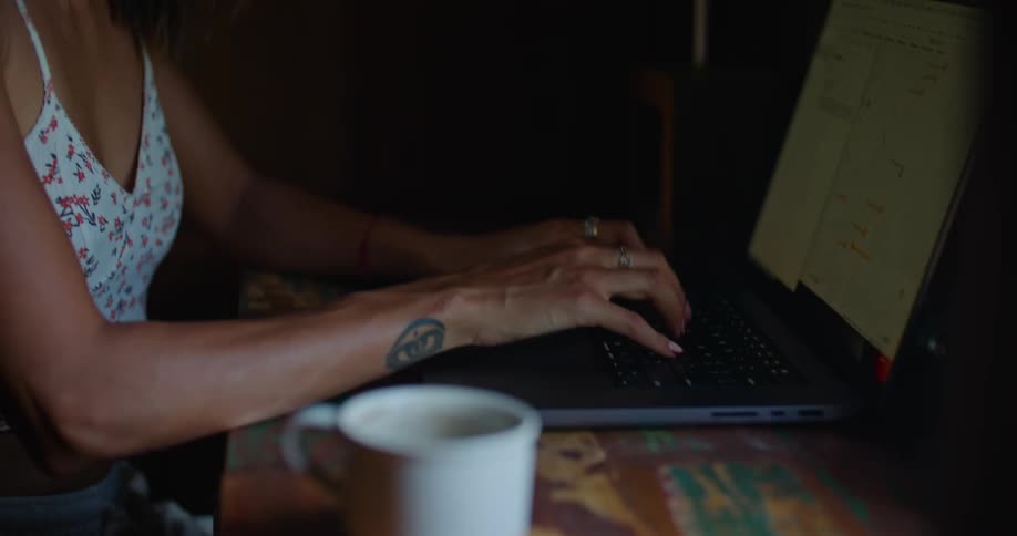 Woman with straight hair earrings printed halter blouse and tattoo types on laptop smiling