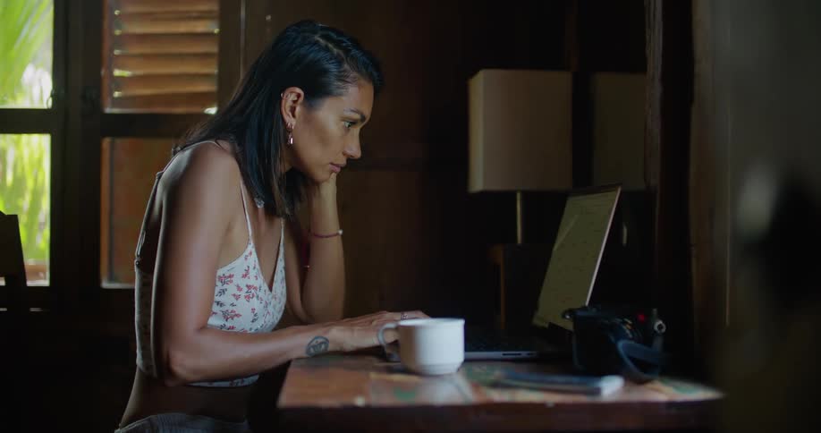 Woman with straight hair earrings printed halter blouse and tattoo types on laptop smiling