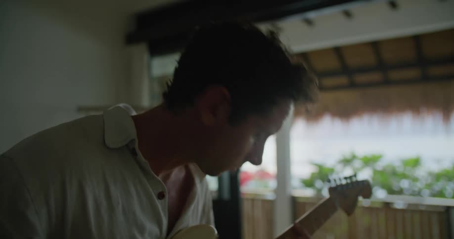 Man with white polo shirt sits on bed playing white electric guitar and looking at laptop
