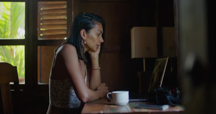 A woman takes a sip of her coffee while talking on video call at her home office