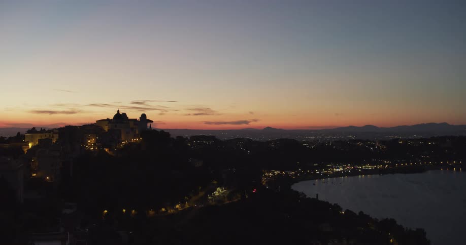 Domed building on mound stands silhouetted against red sky as city lights shine against dark city