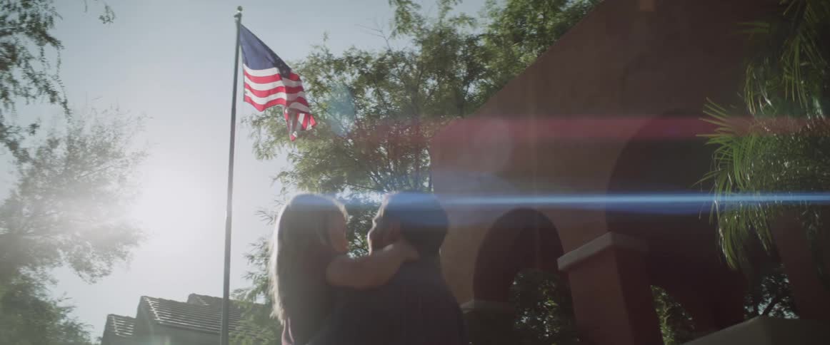 A man holds his daughter while they stand outside and watch an american flag