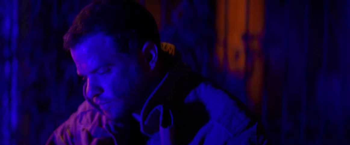A firefighter sitting outdoors removes his helmet while illuminated by the flashing lights of an emergency vehicle at night