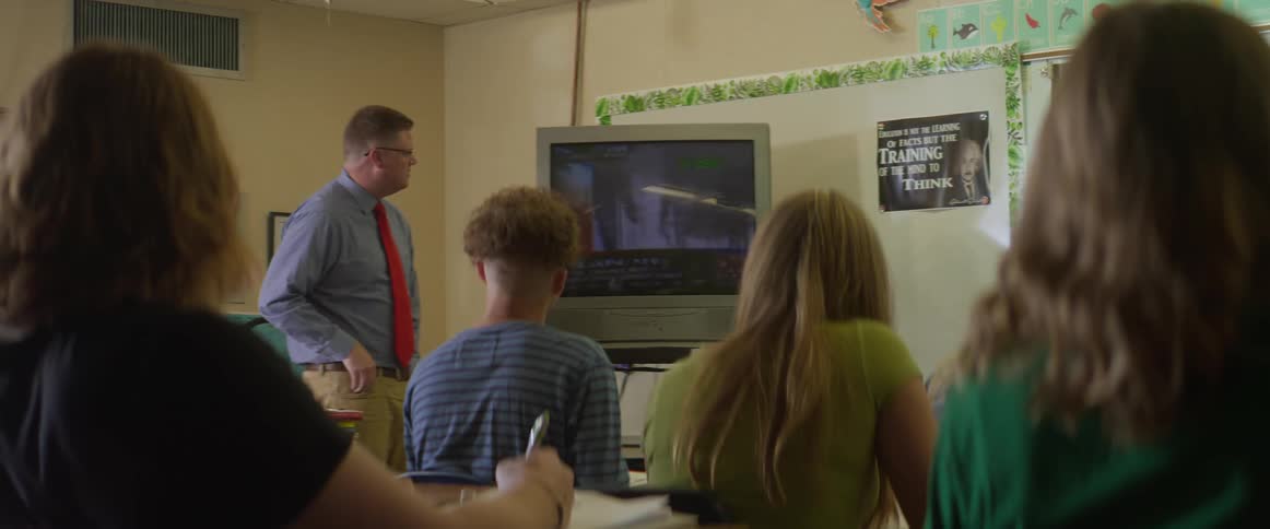 A teacher is standing in his classroom and he turns on the television for his students to watch