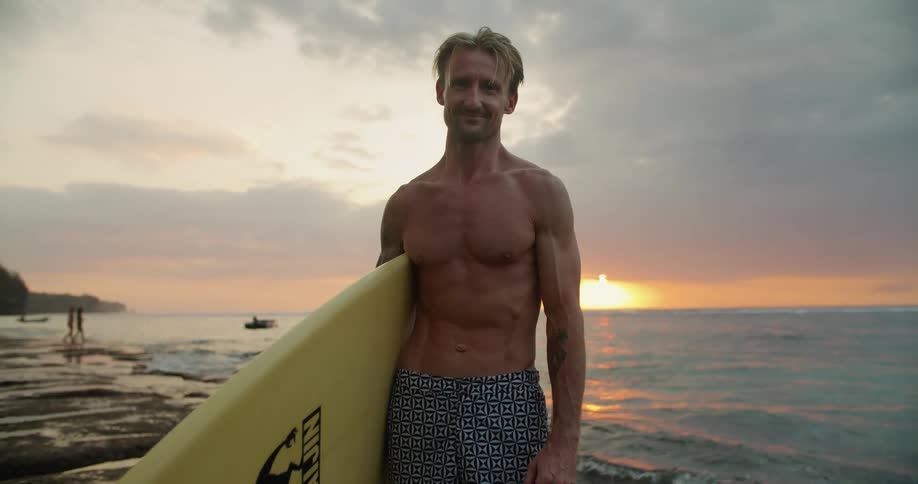A surfer standing on a beach with a surfboard at sunset