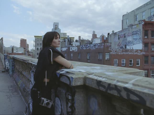 A woman with a camera over her shoulder is leaning on a wall looking at buildings under a cloudy sky