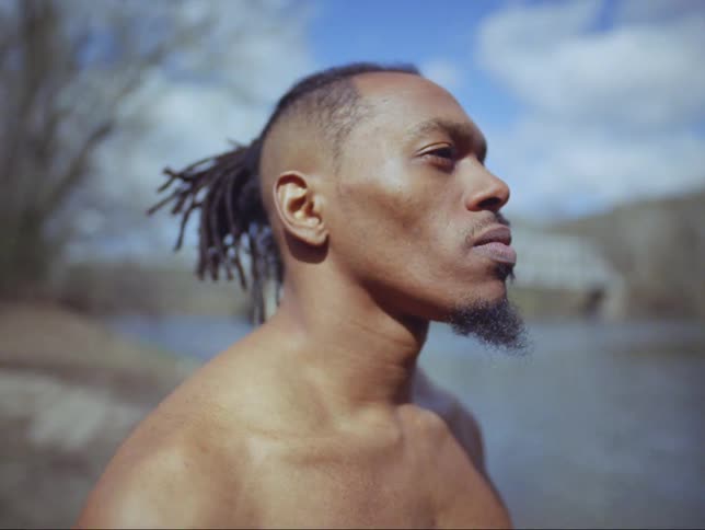 There is a young black man with a bare chest stands in a wooded area near a river and bridge