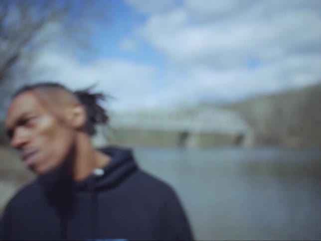 A man is standing along the side of a river near woods with automobiles crossing a bridge in the distance and he turns and walks away