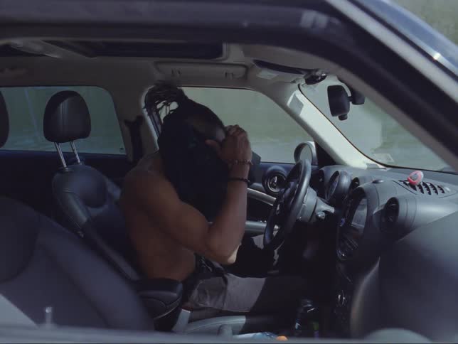 A shirtless black man enters his car and put on his shirt