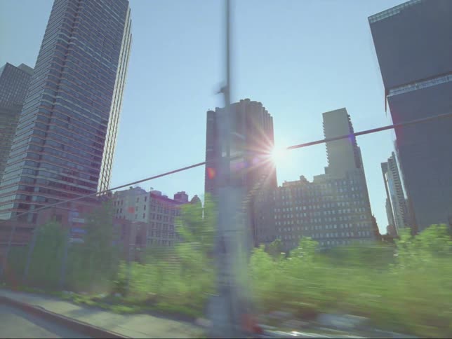 Moving along a chain linked fence tall city buildings are standing in a clear blue sky with the sun shining brightly in the distance