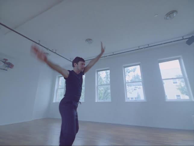 A young white man is dancing inside of a studio brightly lit by sunlight