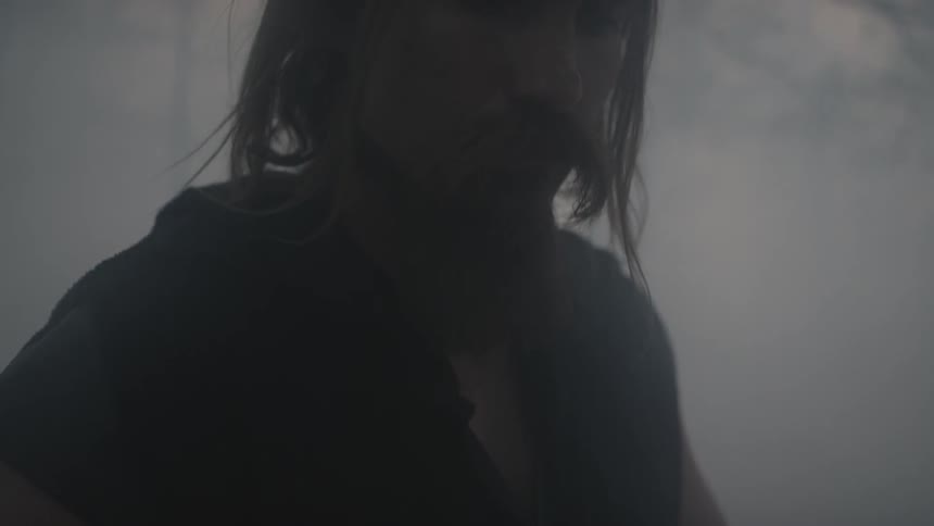 A man stands outside in the mist with his hands resting on a weapon