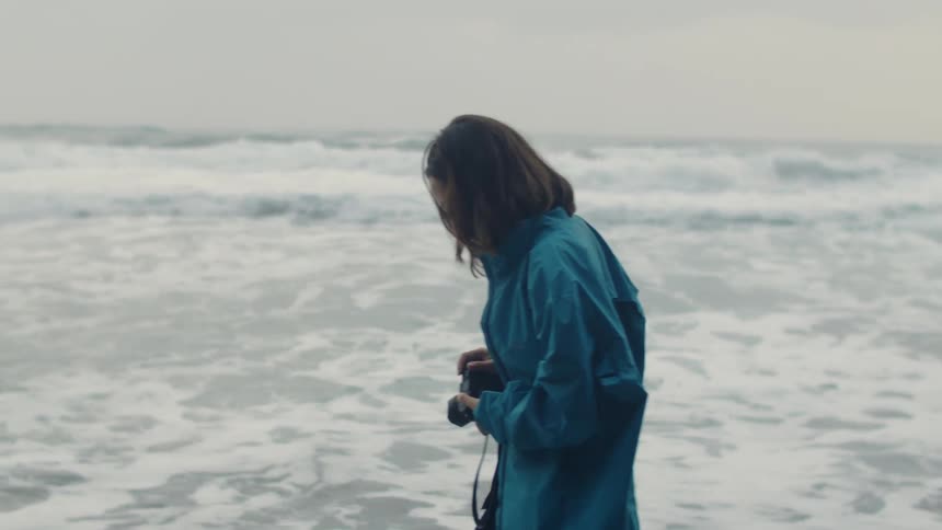 A teenage girl walks along the shore carrying a camera as waves rush in