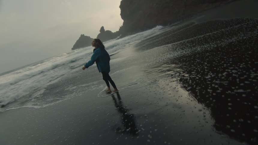 A woman is walking barefoot on a beach and looks up at the sky as she walks through the water