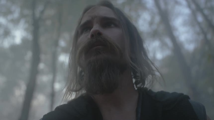 A man with a dirty face and shaggy hair stands still in a forest