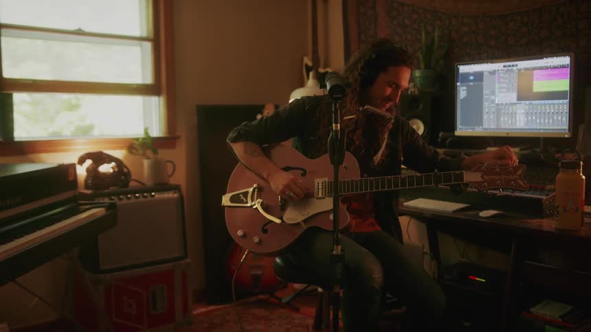 A man is sitting in a chair with a microphone in front of him as he tunes his guitar